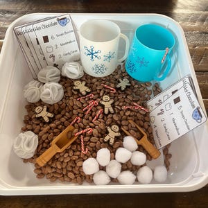 May include: A white sensory bin filled with brown beans, white cotton balls, red and white candy canes, wooden figurines, and two blue mugs. There are also several white cards with the text "Ho! Ho! Hot Chocolate" and a list of ingredients for making hot chocolate.