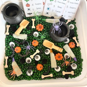 May include: A Halloween-themed sensory bin with a variety of elements. Includes small cauldrons, wooden scoops, and cards that read "Witches Brew". The bin contains green beads, orange pumpkins, spiders, bones, and eyeballs.