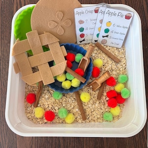 May include: A sensory bin with felt apple pie pieces, pom-poms, wooden scoops, and cinnamon sticks. Includes cards with text: "Apple Crisp" and "Apple Pie" with ingredient lists. The bin is filled with oats.