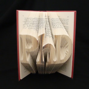 May include: A book with pages folded into the shape of the word "READ" in a 3D style. The book is open and the pages are folded to create the letters. The book is bound in red.