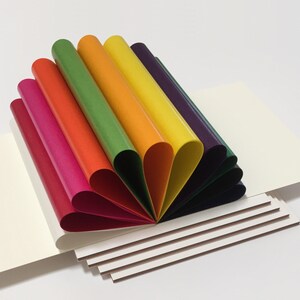 May include: A fan of paper sheets folded in half, creating a book-like shape. The sheets are arranged in a gradient from dark purple to red, with the edges of the sheets visible. The book is resting on a stack of white paper.