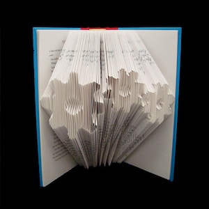 Könnte beinhalten: Ein weißes Buch mit Seiten, die zu einem 3D-Design gefaltet sind und eine Schneeflockenform bilden. Das Buch hat einen blauen Einband und einen roten Rücken.