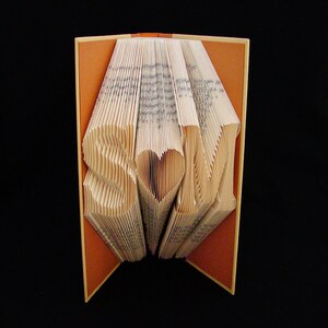 May include: A book art sculpture with the letters "S" and "M" on either side of a heart. The book is folded into a three-dimensional shape and has a brown cover.