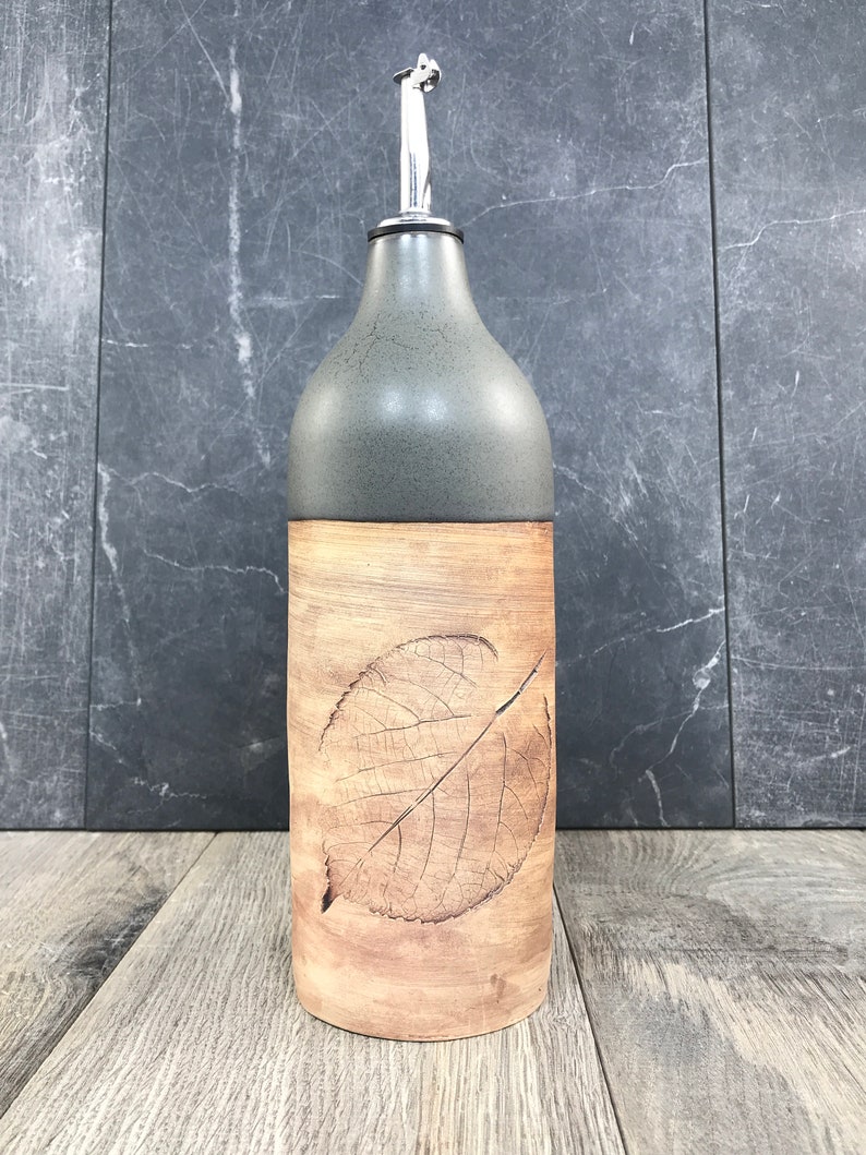 May include: A ceramic olive oil bottle with a gray top and a brown bottom. The bottle has a leaf design embossed on the front.