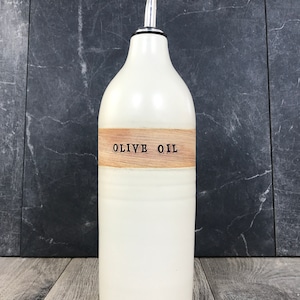K&ouml;nnte beinhalten: Wei&szlig;e Keramik-Oliven&ouml;lflasche mit einem Holzetikett mit der Aufschrift "OLIVE OIL". Die Flasche hat einen silbernen Ausgie&szlig;er.