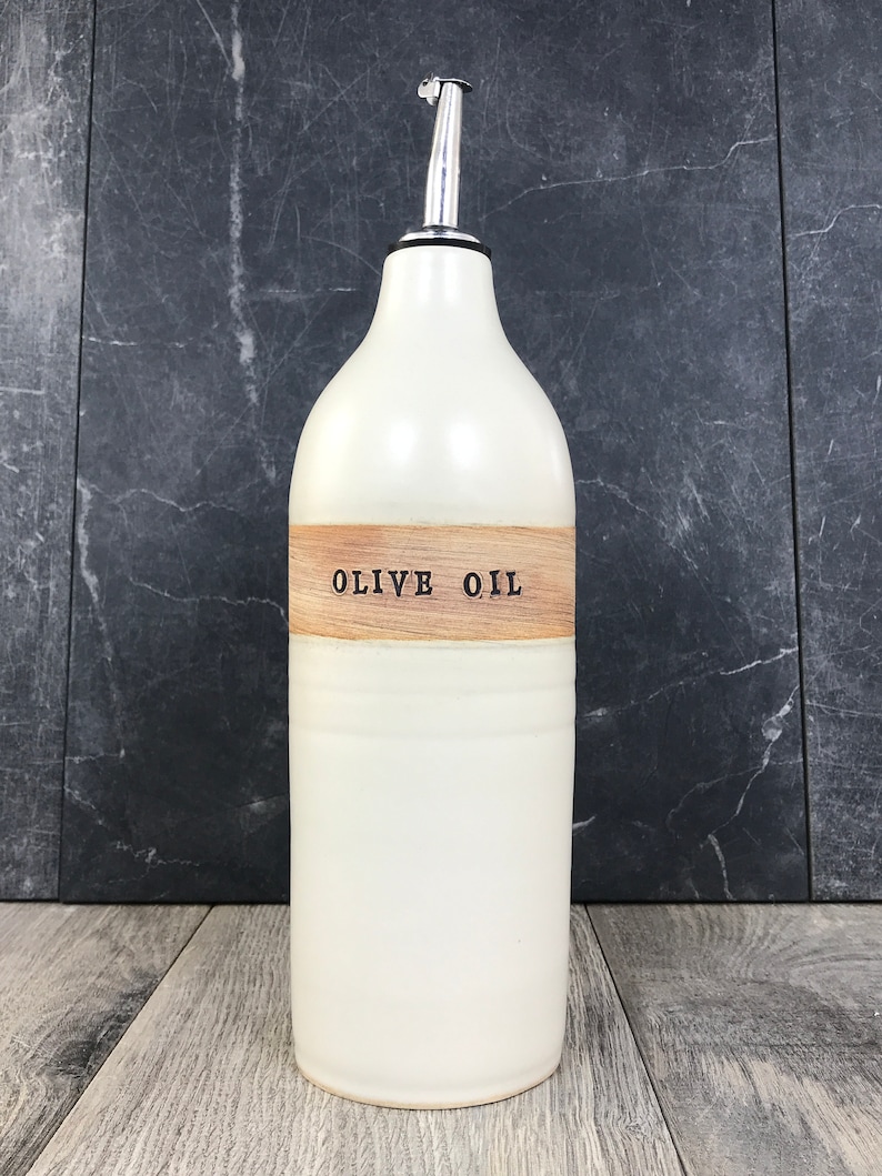 K&ouml;nnte beinhalten: Wei&szlig;e Keramik-Oliven&ouml;lflasche mit einem Holzetikett mit der Aufschrift "OLIVE OIL". Die Flasche hat einen silbernen Ausgie&szlig;er.