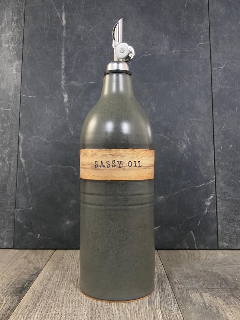 K&ouml;nnte beinhalten: Eine dunkelgraue Keramik-&Ouml;l-Flasche mit einem Holzetikett, auf dem "SASSY OIL" steht. Die Flasche hat einen silbernen Ausgie&szlig;er und steht auf einer Holzoberfl&auml;che.