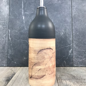 May include: A black and brown ceramic oil and vinegar bottle with a leaf design. The bottle has a silver pour spout.