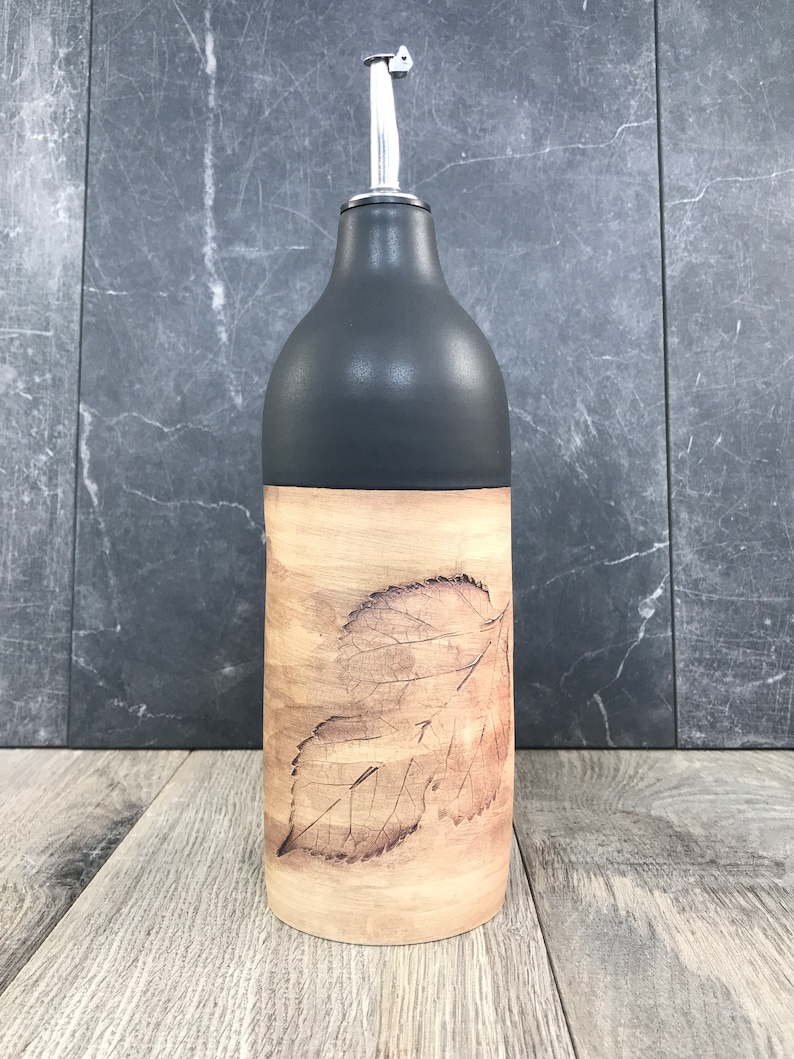 May include: A black and brown ceramic oil and vinegar bottle with a leaf design. The bottle has a silver pour spout.