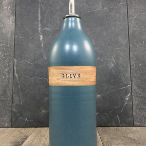 K&ouml;nnte beinhalten: Eine blaue Keramik-Oliven&ouml;lflasche mit einem Holzetikett, auf dem "OLIVE" steht. Die Flasche hat einen silbernen Ausgie&szlig;er.