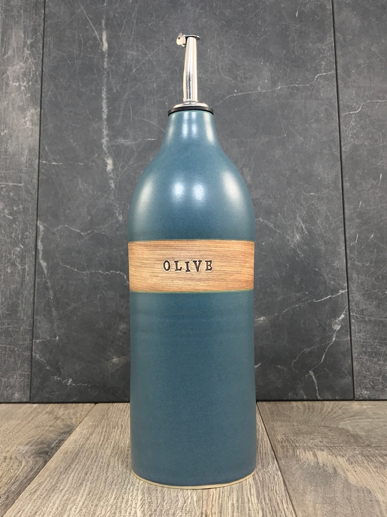 K&ouml;nnte beinhalten: Eine blaue Keramik-Oliven&ouml;lflasche mit einem Holzetikett, auf dem "OLIVE" steht. Die Flasche hat einen silbernen Ausgie&szlig;er.
