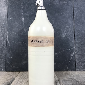 K&ouml;nnte beinhalten: Wei&szlig;e Keramikflasche mit einem Holzetikett mit der Aufschrift "GARLIC OIL". Die Flasche hat einen silbernen Pumpspender.