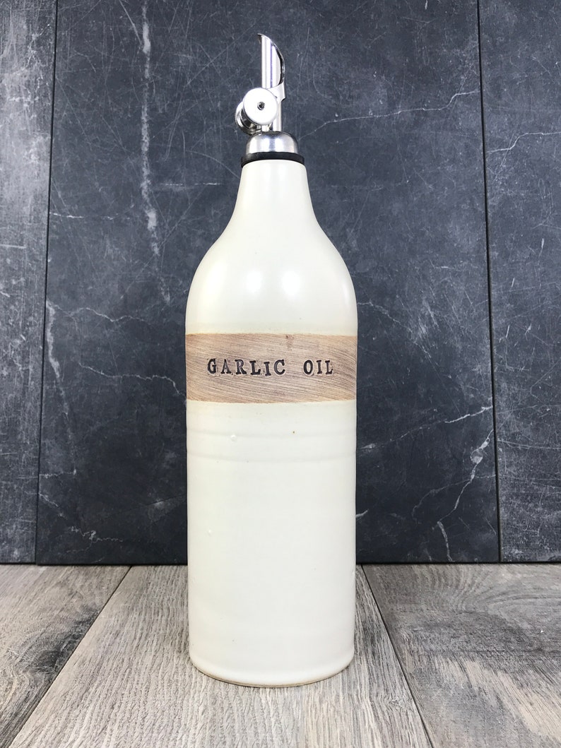 K&ouml;nnte beinhalten: Wei&szlig;e Keramikflasche mit einem Holzetikett mit der Aufschrift "GARLIC OIL". Die Flasche hat einen silbernen Pumpspender.