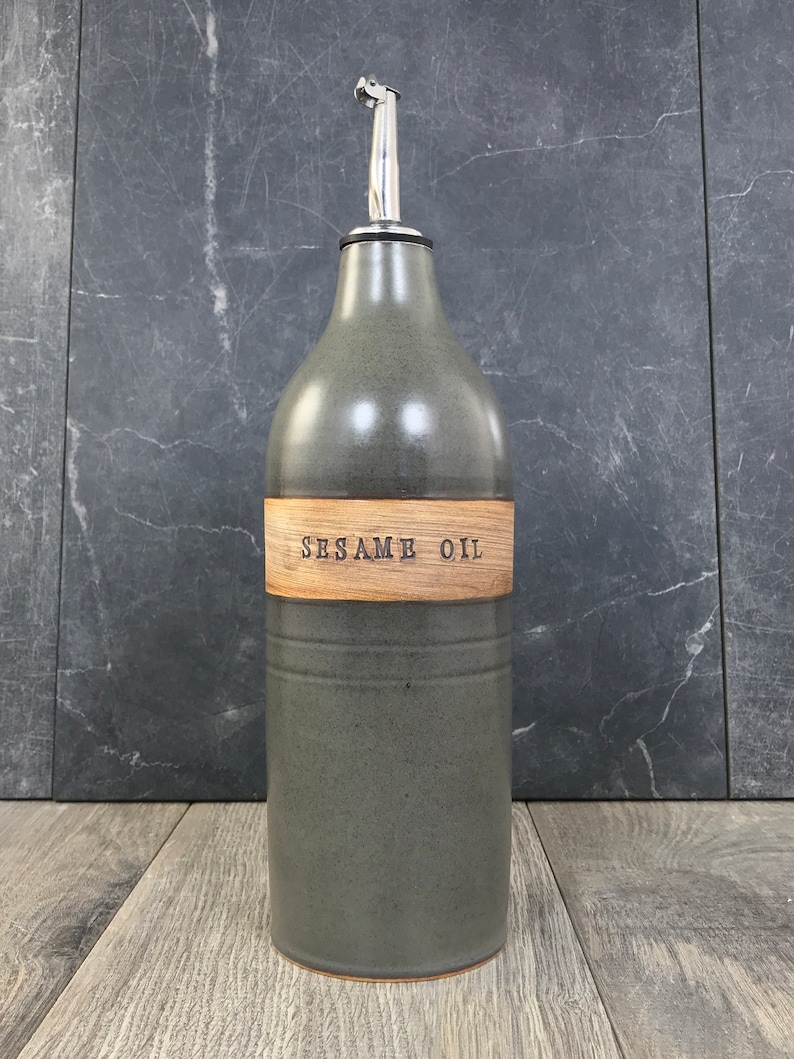 K&ouml;nnte beinhalten: Eine graue Keramik-&Ouml;l-Flasche mit einem Holzetikett, auf dem "SESAME OIL" steht. Die Flasche hat einen silbernen Ausgie&szlig;er und steht auf einer Holzoberfl&auml;che.