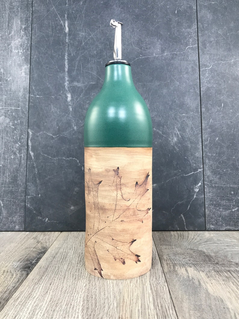 May include: A green ceramic olive oil bottle with a silver pour spout and a brown leaf design.