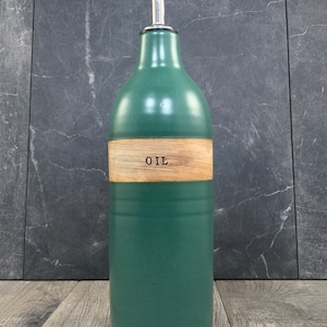 K&ouml;nnte beinhalten: Eine gr&uuml;ne Keramik-&Ouml;l-Flasche mit einem Holzetikett, auf dem "OIL" steht. Die Flasche hat einen silbernen Ausgie&szlig;er.