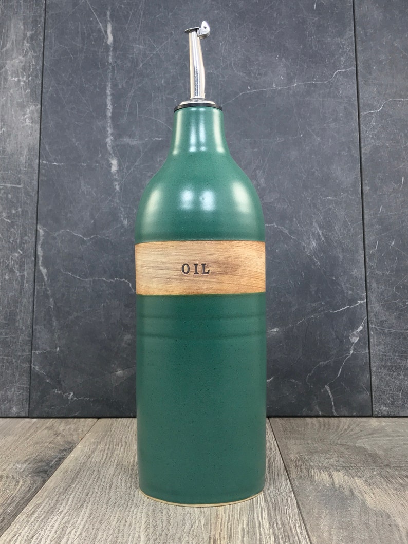 K&ouml;nnte beinhalten: Eine gr&uuml;ne Keramik-&Ouml;l-Flasche mit einem Holzetikett, auf dem "OIL" steht. Die Flasche hat einen silbernen Ausgie&szlig;er.
