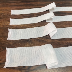 May include: Four white elastic bands of varying widths, lying flat on a wooden surface. The bands are made of a soft, stretchy material and are perfect for sewing projects.