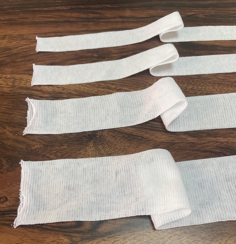 May include: Four white elastic bands of varying widths, lying flat on a wooden surface. The bands are made of a soft, stretchy material and are perfect for sewing projects.