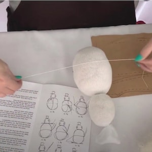 May include: A person is sewing a white fabric ball with a white thread. The ball is on a table with a brown piece of fabric and a set of instructions. The instructions show how to sew a stuffed toy.