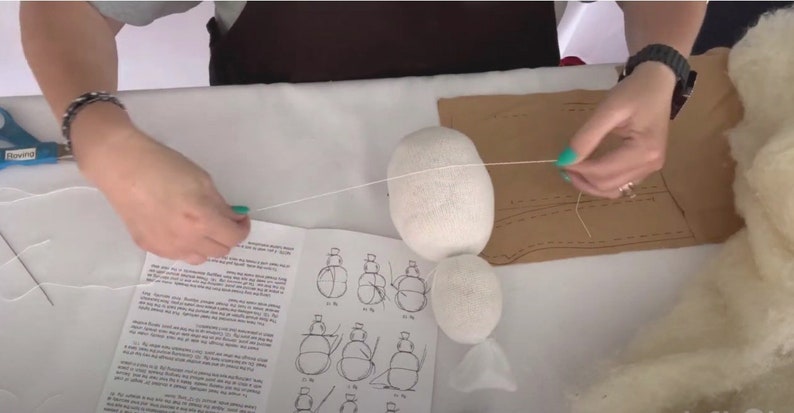 May include: A person is sewing a white fabric ball with a white thread. The ball is on a table with a brown piece of fabric and a set of instructions. The instructions show how to sew a stuffed toy.
