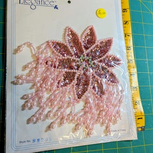 May include: A pink floral applique with sequins and pearl bead fringe, packaged on a card. The applique has a central cluster of iridescent rhinestones. The card reads "Elegance" and "Made in China". Style No. SM205.