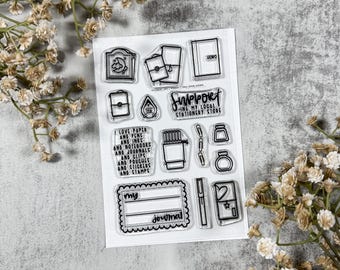 Op de afbeelding: Transparante stempelset met verschillende ontwerpen met een briefpapier thema, waaronder notitieboekjes en stempels, en de tekst "Support My Local Stationery Store". De set bevat ook de tekst "I love paper and pens and inks and notebooks and journals and clips and pouches and stickers and stamps."