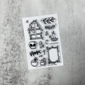 May include: Clear stamp set featuring various mystical and witchy designs. Includes a skull, window, candle, crystals, and the word "Magic". Also includes a key, potion bottles, and a decorative frame. The stamps are black and white.