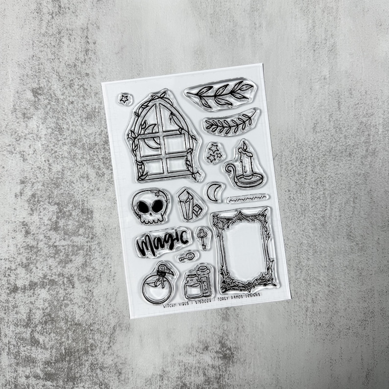 May include: Clear stamp set featuring various mystical and witchy designs. Includes a skull, window, candle, crystals, and the word "Magic". Also includes a key, potion bottles, and a decorative frame. The stamps are black and white.