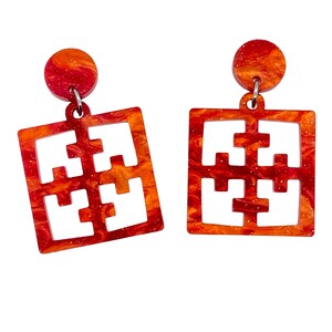 May include: A pair of orange and red marbled acrylic earrings. Each earring features a small circular stud connected to a square pendant with a cut-out puzzle-like design. The earrings have a shiny, speckled finish.