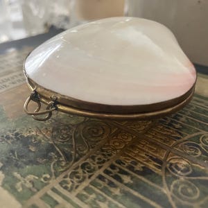 Könnte beinhalten: Kleine Schmuckschatulle in Muschelform mit perlweißem Deckel und einem dünnen, goldfarbenen Metallrahmen. Die Schatulle hat ein Scharnier und eine kleine Schlaufe zum Anbringen einer Kette. Die Schatulle liegt auf einer dekorativen Oberfläche.