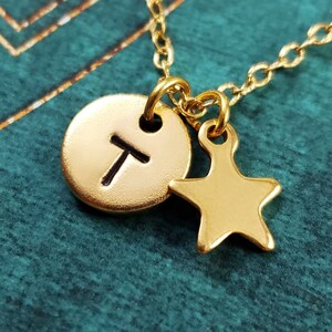 Könnte beinhalten: Goldkette mit einem kleinen runden Anhänger mit dem Buchstaben "T" und einem kleinen goldenen Sternanhänger.