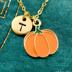 May include: A gold chain necklace with a small gold circle charm with the letter "T" and a pumpkin charm. The pumpkin charm is orange with a green stem.