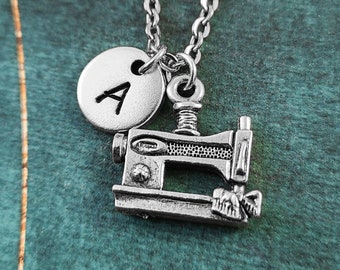 May include: Silver necklace featuring a sewing machine charm and a silver disc charm with the letter A. The necklace is displayed against a green background.