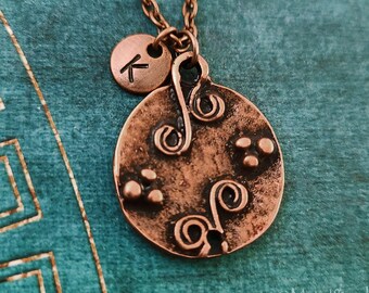 May include: A copper necklace with a round pendant featuring a stylized design of swirls and dots. A smaller round charm with the letter "K" is attached to the pendant.