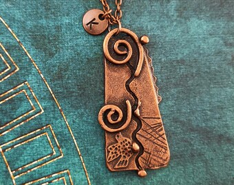 May include: A copper necklace with a pendant featuring a stylized bird design and intricate swirls. The pendant is attached to a copper chain with a small charm that says "K".
