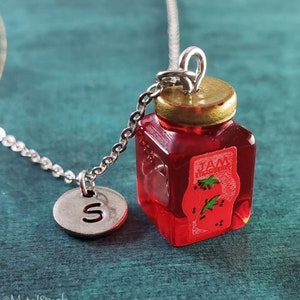 May include: A silver necklace with a miniature red jam jar pendant, a gold-coloured lid, and a silver charm with the letter "S". The jam jar has a label that reads "JAM" with a strawberry illustration.