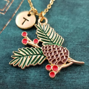 May include: Gold necklace featuring a pine cone, pine branches, and red berry charm. The necklace also has a gold initial charm with the letter "T".