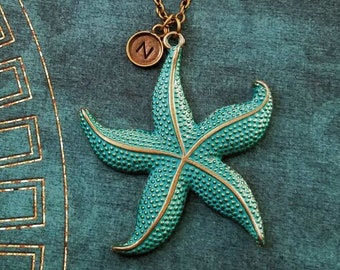 May include: A turquoise blue starfish pendant necklace with a gold-toned chain and a small, round, brown charm with the letter "N" on it.