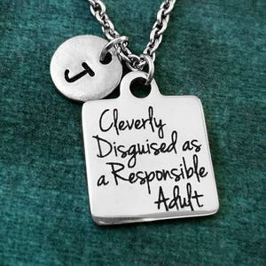 May include: Silver necklace featuring a square pendant engraved with "Cleverly Disguised as a Responsible Adult." A small, round charm with the letter "J" is also present. The chain has a linked pattern.