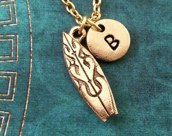 May include: Gold necklace with a surfboard charm and a letter 'B' charm. The necklace is on a teal background.