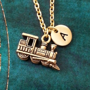 Peut inclure: Collier en or avec un petit pendentif en forme de train et un pendentif rond avec la lettre A. Le collier est sur un fond bleu sarcelle.