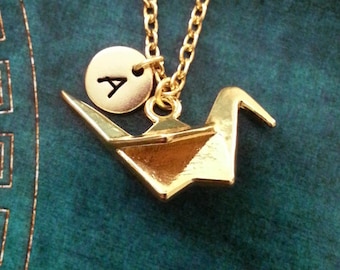 May include: Gold origami crane necklace with a gold initial charm. The initial charm is a circle with the letter 'A' in the center.