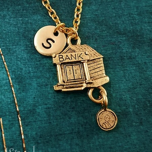 May include: Gold necklace with a charm of a small building with the word "BANK" above the entrance. The necklace also has a charm with the letter "S" and a small round charm with a design.