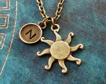 May include: A gold necklace with a sun charm and a small charm with the letter N. The necklace is on a blue background.