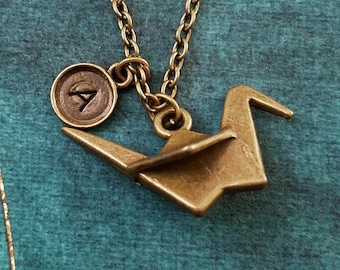May include: A gold-toned necklace with a small origami crane charm and a circular pendant with the letter 'A' inscribed on it.