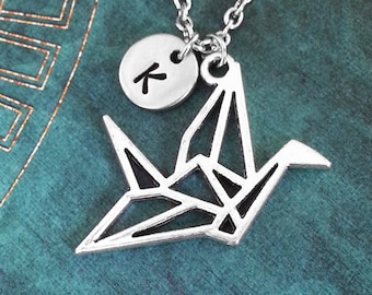 May include: Silver-toned necklace featuring a geometric origami crane pendant and a small circular charm with the letter "K". The chain and charms are displayed against a teal background.