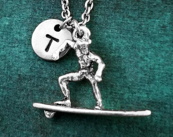 May include: Silver necklace with a surfer charm and a round charm with the letter 'T' on it. The necklace is on a green background.