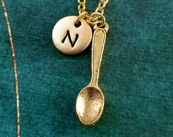 May include: Gold necklace with a small spoon charm and a round charm with the letter N. The necklace is on a green background.