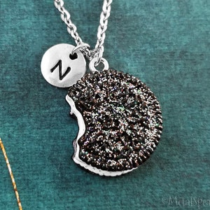 May include: A silver necklace with a charm of a chocolate chip cookie with a bite taken out of it. The cookie charm is covered in glitter. The necklace also has a silver charm with the letter "N" on it.